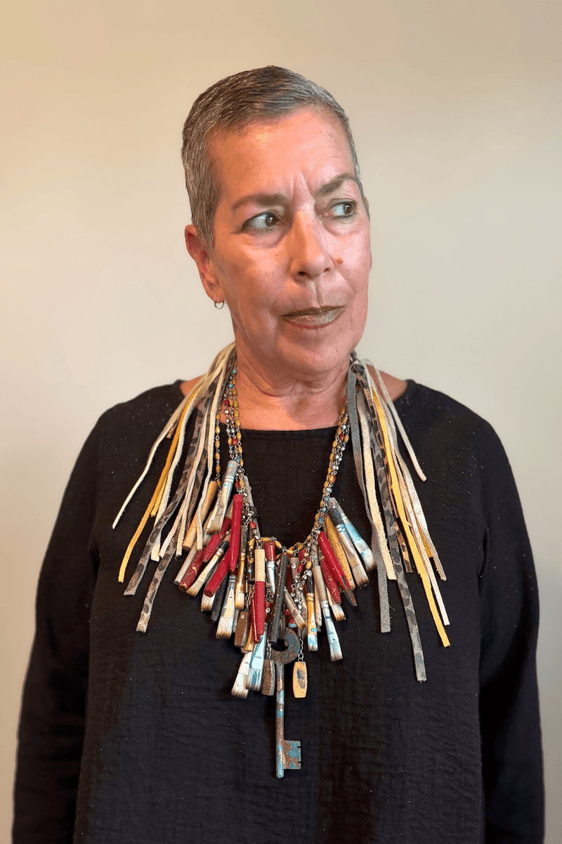 A Rare Bird Painter's Palette Layered Necklace, a wide bib-style statement necklace with layered paint-marked brushes, colorful beads, chains, dangling artist-inspired objects, and long mixed leather fringe ties.