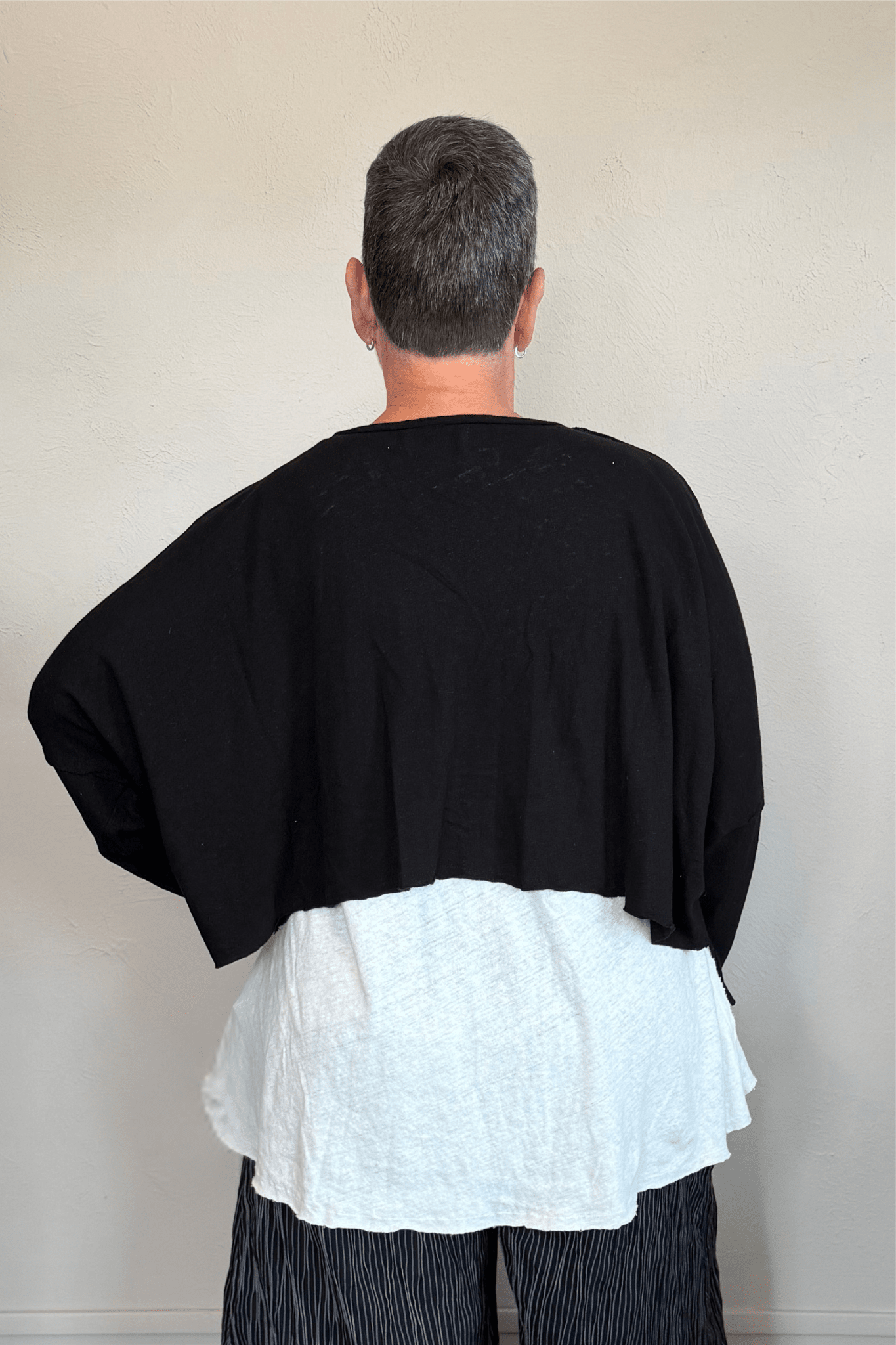 Cropped knit cardigan jacket shown in black and white, with a relaxed boxy shape, dropped shoulders, long sleeves, assorted front buttons, curved seam detailing, raw-edge finishes, and hanging cord accents.