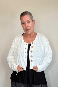 Cropped knit cardigan jacket shown in black and white, with a relaxed boxy shape, dropped shoulders, long sleeves, assorted front buttons, curved seam detailing, raw-edge finishes, and hanging cord accents.
