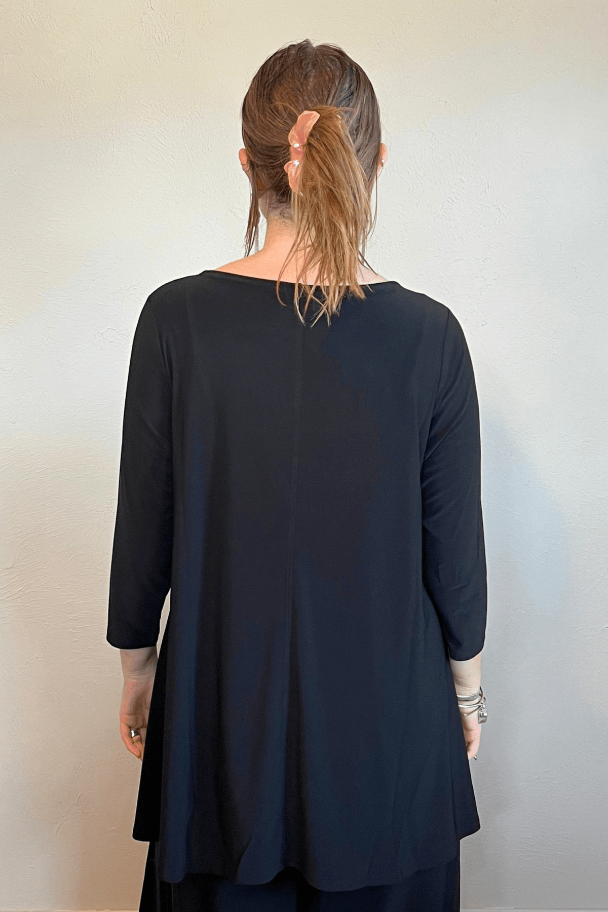 Model wearing the black Kozan Dima tunic with a boat neckline, three-quarter sleeves, and an asymmetrical draped hem, shown from front and back.