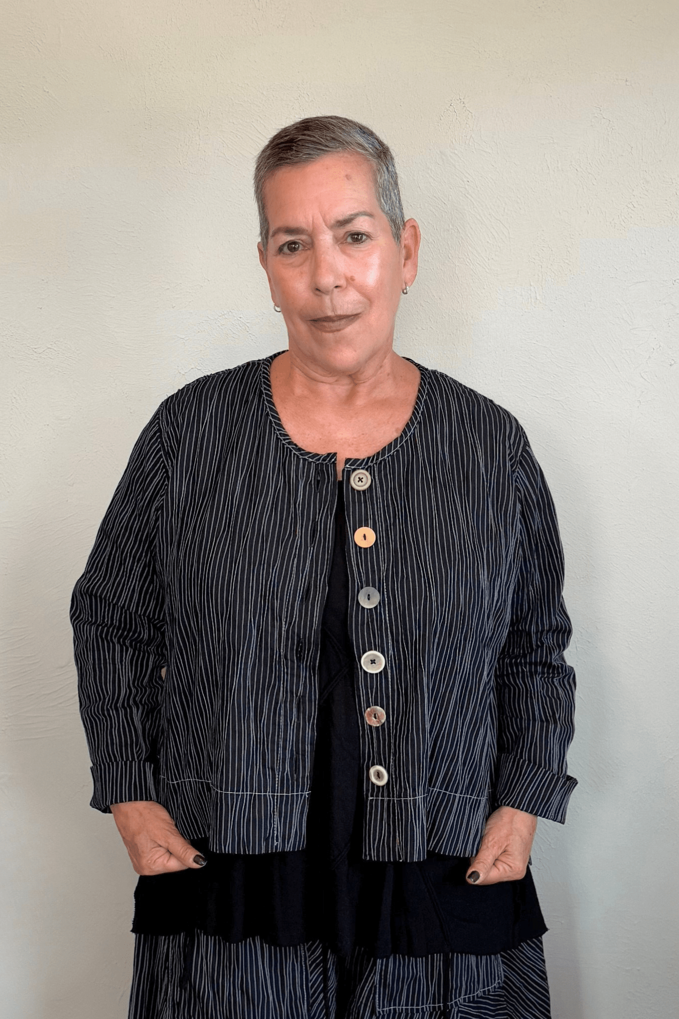 Black cropped button-front jacket with fine vertical white striping, collarless round neckline, long sleeves with turned cuffs, assorted buttons, contrast topstitching, and a relaxed boxy silhouette.