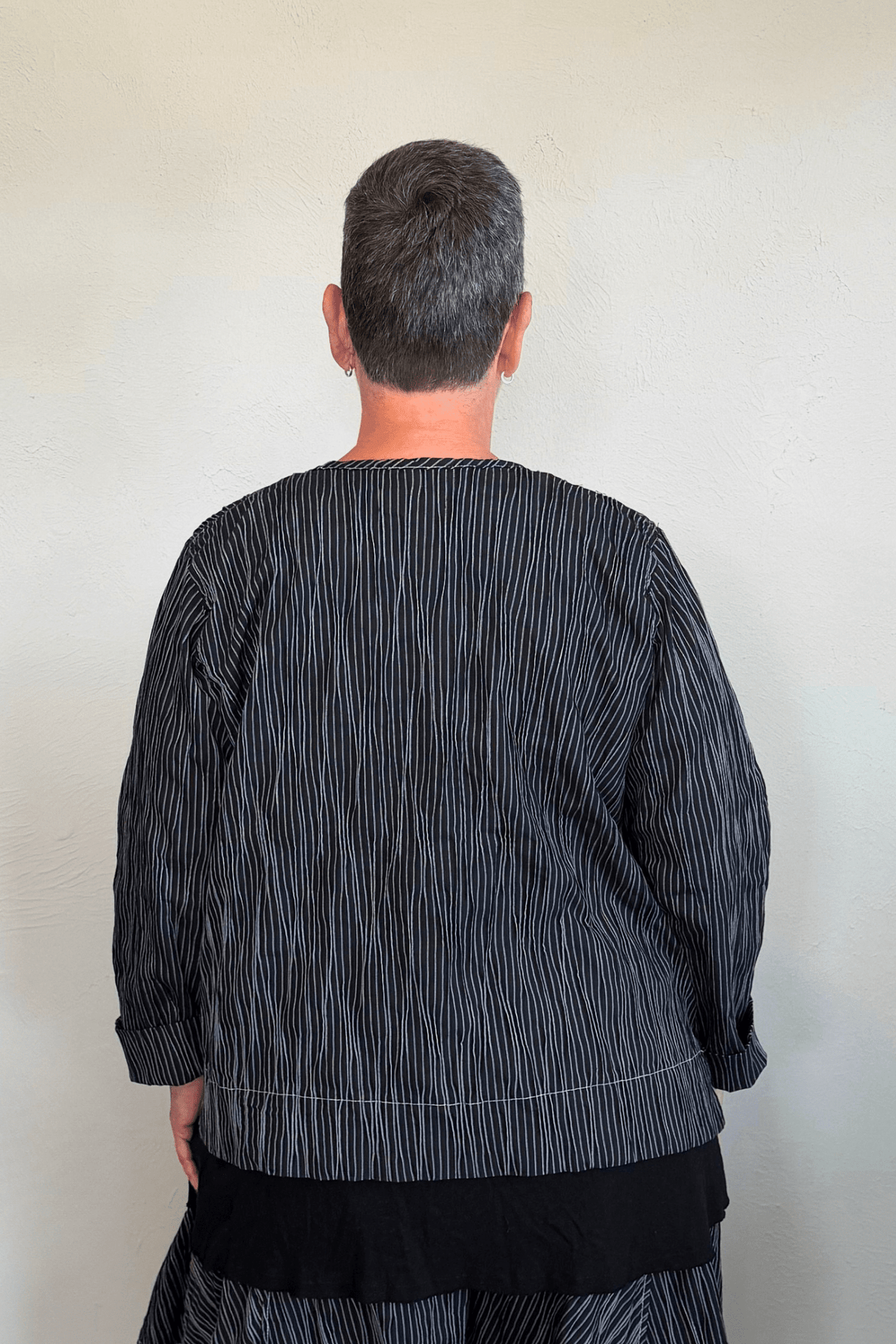 Black cropped button-front jacket with fine vertical white striping, collarless round neckline, long sleeves with turned cuffs, assorted buttons, contrast topstitching, and a relaxed boxy silhouette.