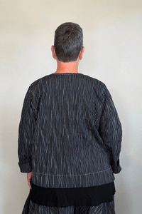 Black cropped button-front jacket with fine vertical white striping, collarless round neckline, long sleeves with turned cuffs, assorted buttons, contrast topstitching, and a relaxed boxy silhouette.