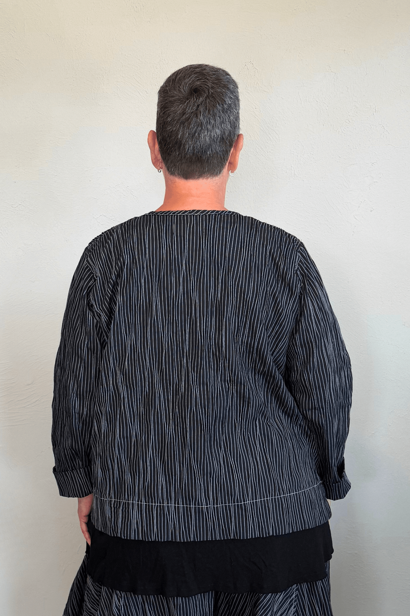 Black cropped button-front jacket with fine vertical white striping, collarless round neckline, long sleeves with turned cuffs, assorted buttons, contrast topstitching, and a relaxed boxy silhouette.