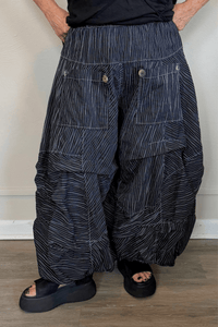 Black full pleat pants with fine vertical white striping, a wide elastic waistband, oversized front patch pockets with button accents, layered asymmetrical panels, and a voluminous balloon-like wide leg.