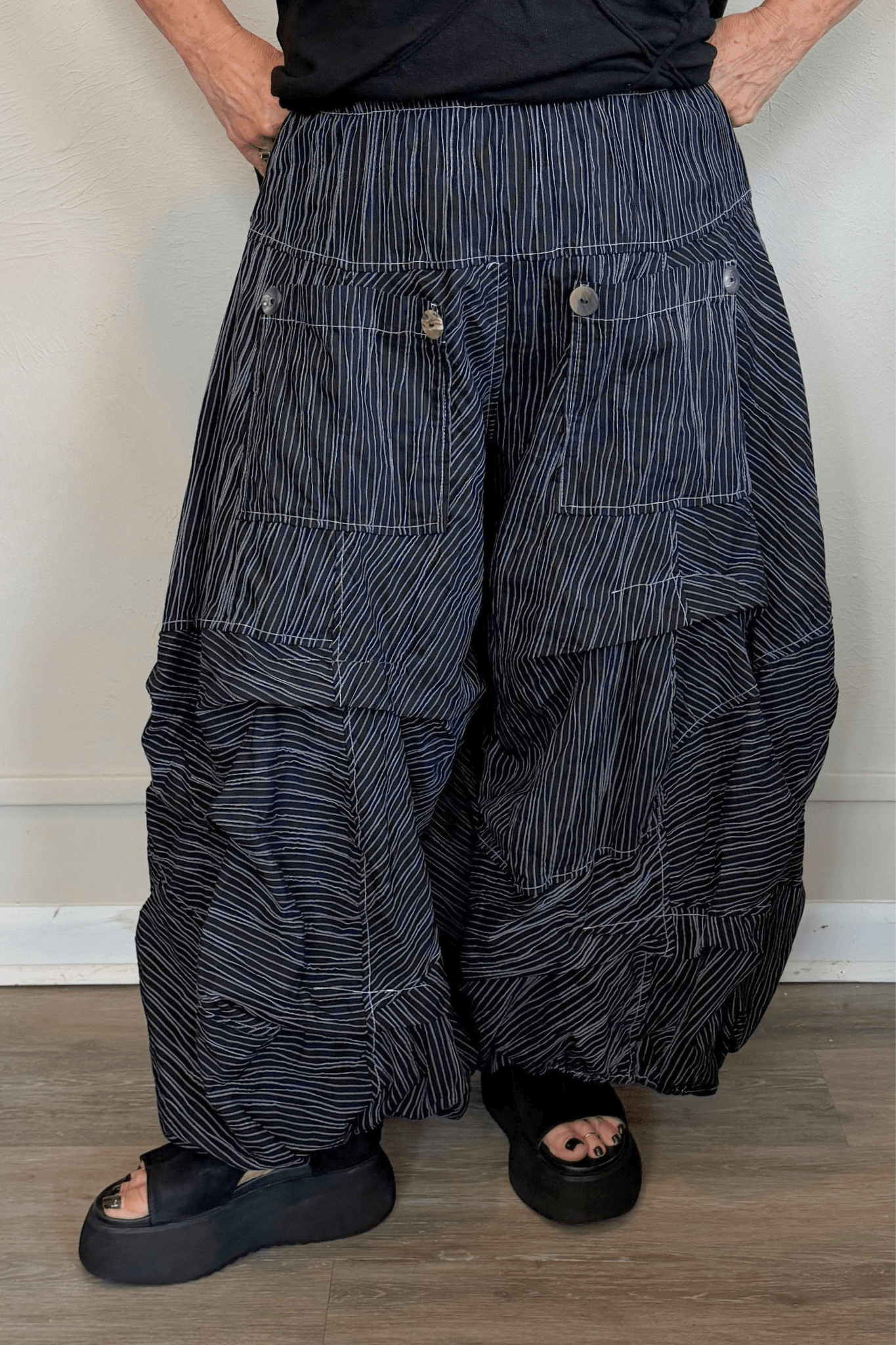 Black full pleat pants with fine vertical white striping, a wide elastic waistband, oversized front patch pockets with button accents, layered asymmetrical panels, and a voluminous balloon-like wide leg.