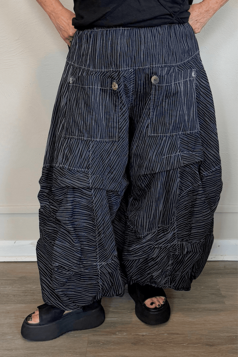 Black full pleat pants with fine vertical white striping, a wide elastic waistband, oversized front patch pockets with button accents, layered asymmetrical panels, and a voluminous balloon-like wide leg.