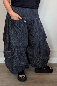 Black full pleat pants with fine vertical white striping, a wide elastic waistband, oversized front patch pockets with button accents, layered asymmetrical panels, and a voluminous balloon-like wide leg.