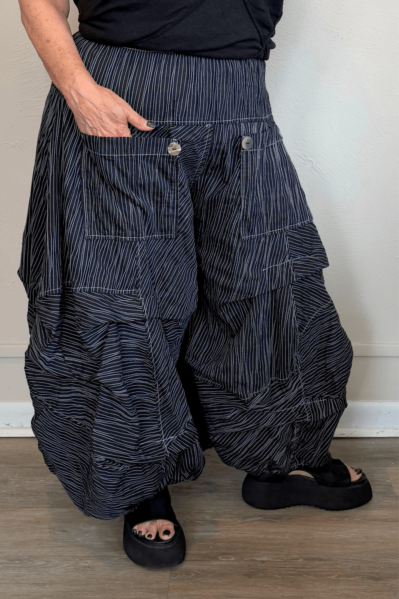 Black full pleat pants with fine vertical white striping, a wide elastic waistband, oversized front patch pockets with button accents, layered asymmetrical panels, and a voluminous balloon-like wide leg.