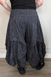 Black full pleat pants with fine vertical white striping, a wide elastic waistband, oversized front patch pockets with button accents, layered asymmetrical panels, and a voluminous balloon-like wide leg.