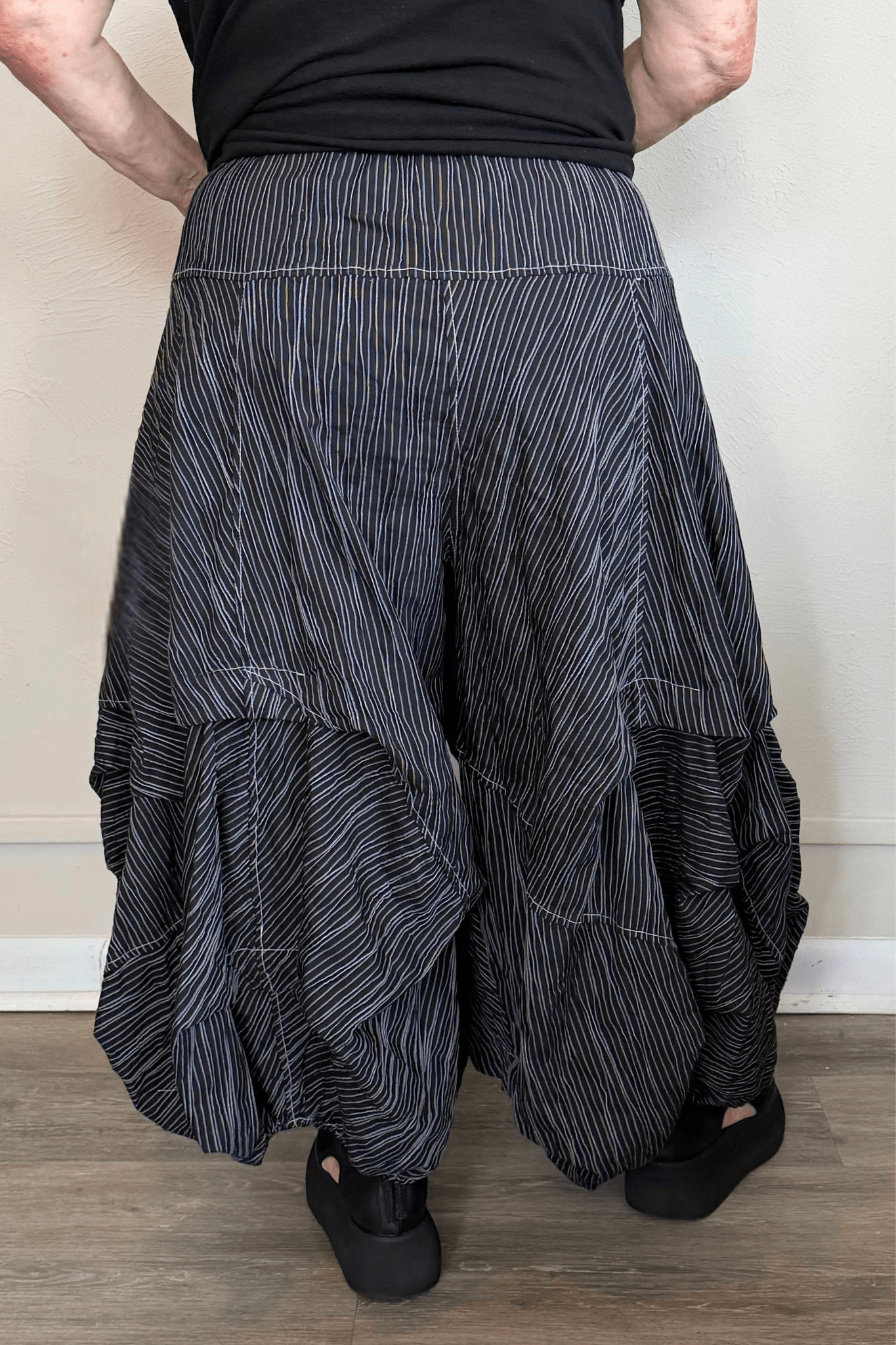 Black full pleat pants with fine vertical white striping, a wide elastic waistband, oversized front patch pockets with button accents, layered asymmetrical panels, and a voluminous balloon-like wide leg.