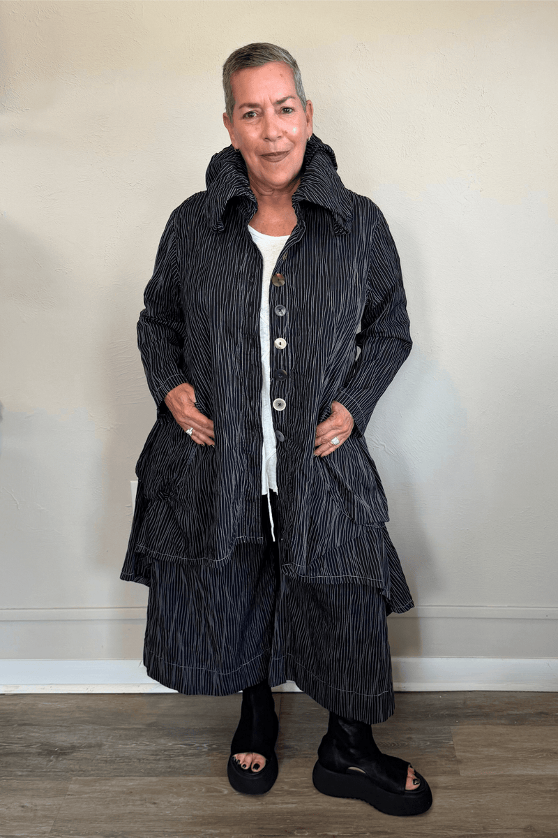 Black long coat with fine vertical white striping, oversized sculptural collar, mixed front buttons, large patch pockets, long sleeves, and an asymmetrical high-low hem with a longer rounded back.