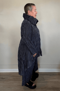 Black long coat with fine vertical white striping, oversized sculptural collar, mixed front buttons, large patch pockets, long sleeves, and an asymmetrical high-low hem with a longer rounded back.