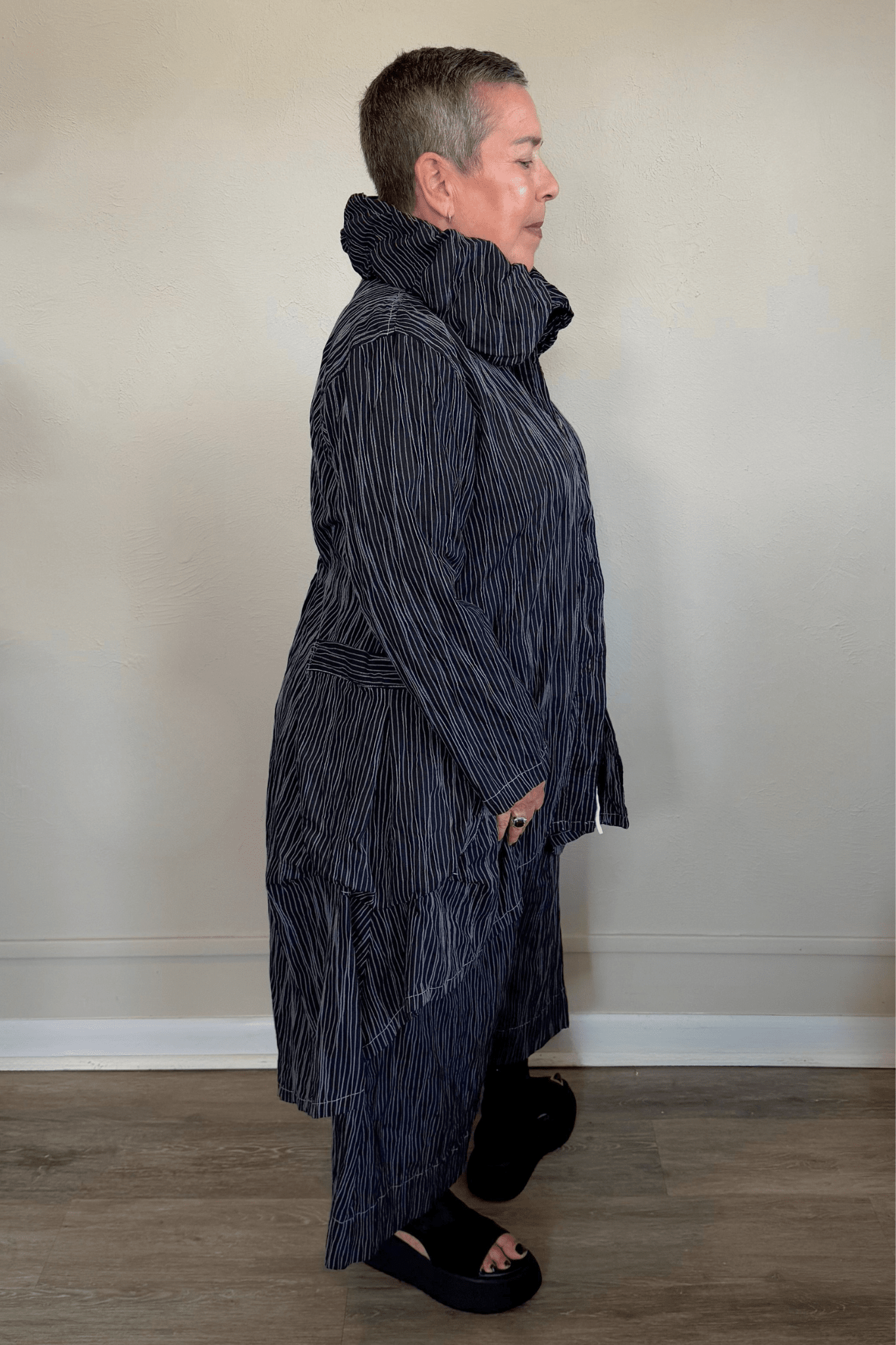 Black long coat with fine vertical white striping, oversized sculptural collar, mixed front buttons, large patch pockets, long sleeves, and an asymmetrical high-low hem with a longer rounded back.