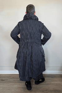 Black long coat with fine vertical white striping, oversized sculptural collar, mixed front buttons, large patch pockets, long sleeves, and an asymmetrical high-low hem with a longer rounded back.