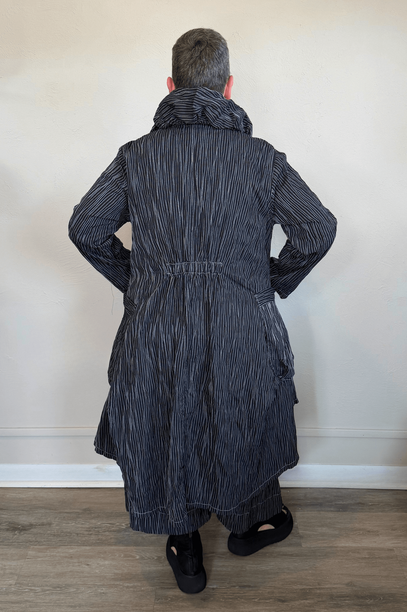 Black long coat with fine vertical white striping, oversized sculptural collar, mixed front buttons, large patch pockets, long sleeves, and an asymmetrical high-low hem with a longer rounded back.