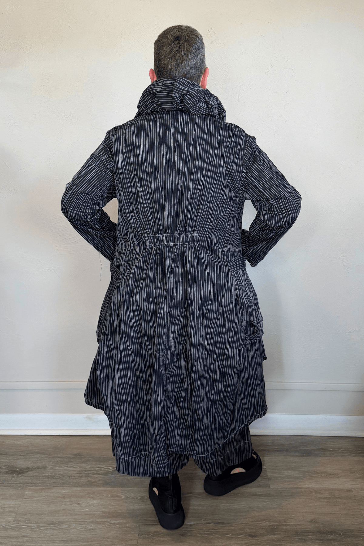 Black long coat with fine vertical white striping, oversized sculptural collar, mixed front buttons, large patch pockets, long sleeves, and an asymmetrical high-low hem with a longer rounded back.