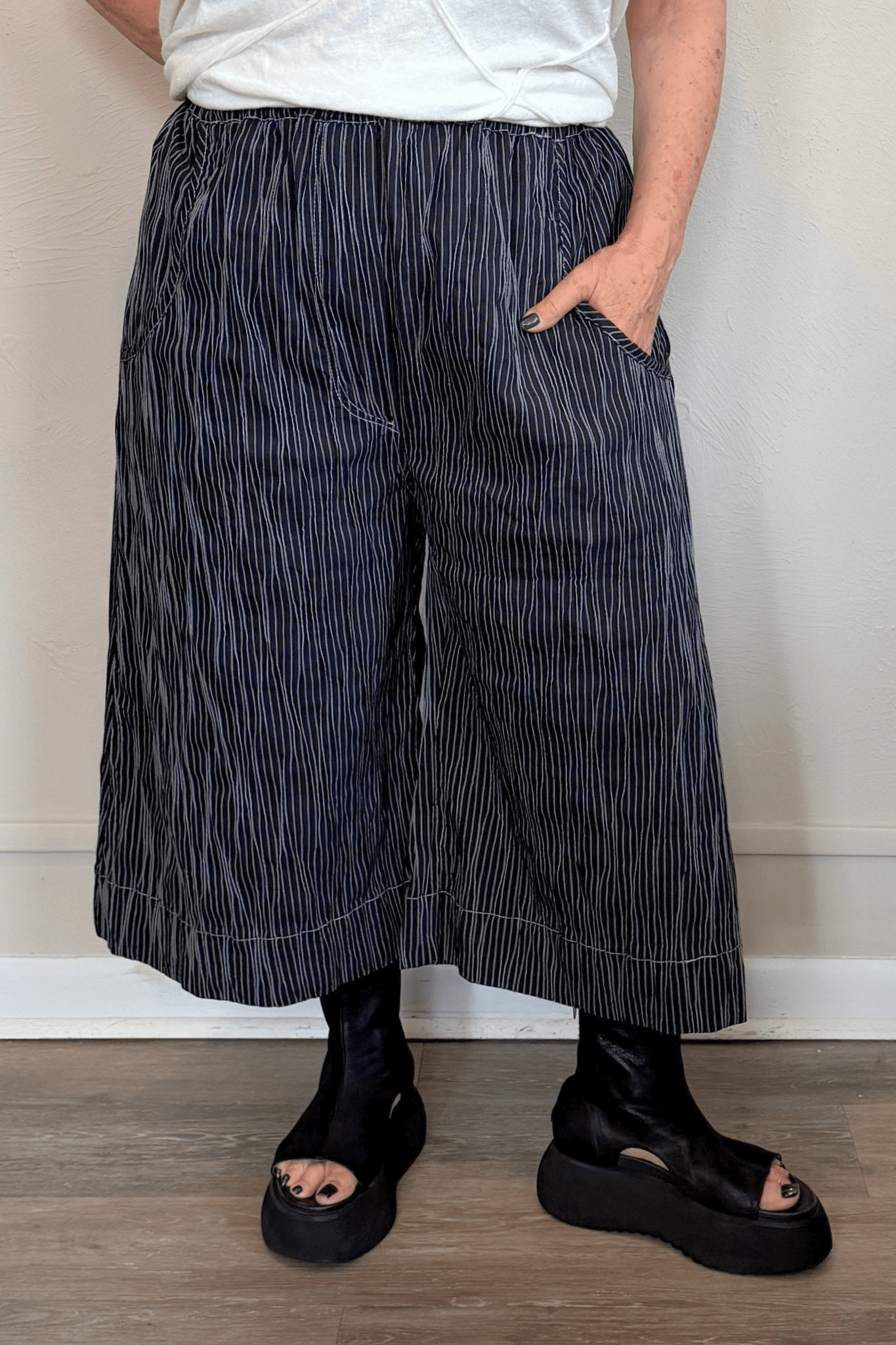 Black cropped wide-leg pants with fine vertical white striping, pull-on elastic waistband, angled side pockets, and contrast topstitching at the hem and pocket edges.