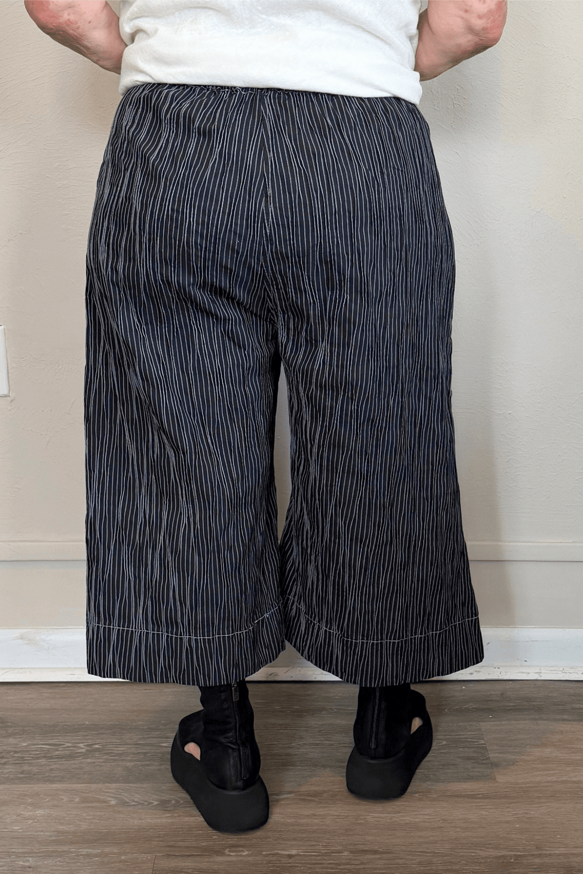 Black cropped wide-leg pants with fine vertical white striping, pull-on elastic waistband, angled side pockets, and contrast topstitching at the hem and pocket edges.