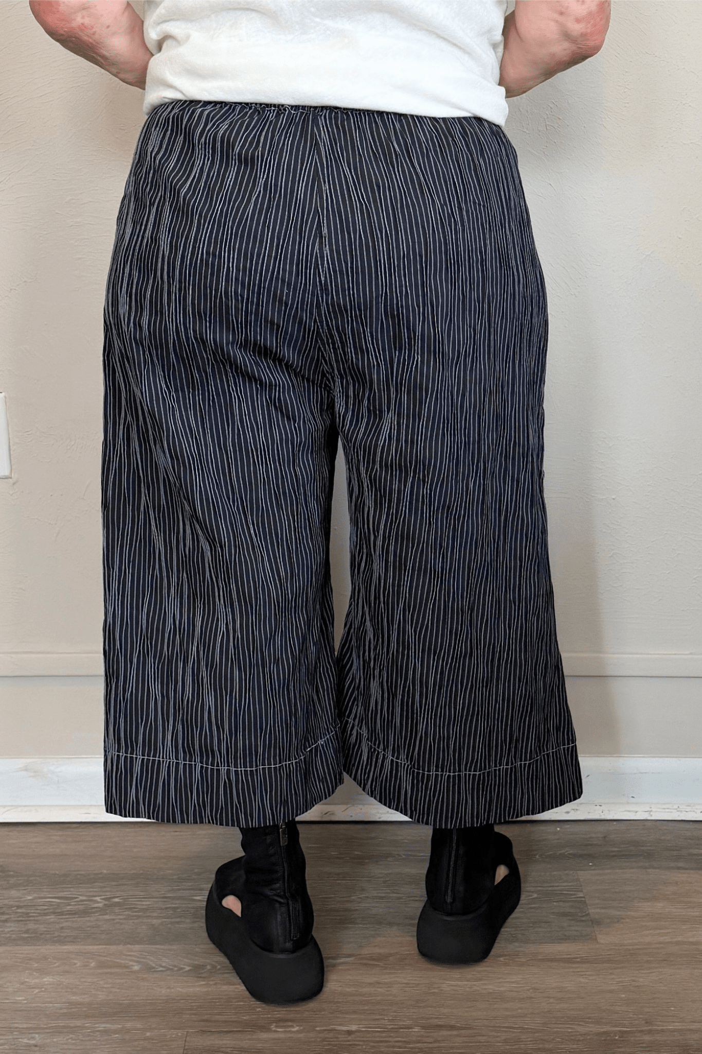 Black cropped wide-leg pants with fine vertical white striping, pull-on elastic waistband, angled side pockets, and contrast topstitching at the hem and pocket edges.