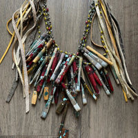 A Rare Bird Painter's Palette Layered Necklace, a wide bib-style statement necklace with layered paint-marked brushes, colorful beads, chains, dangling artist-inspired objects, and long mixed leather fringe ties.