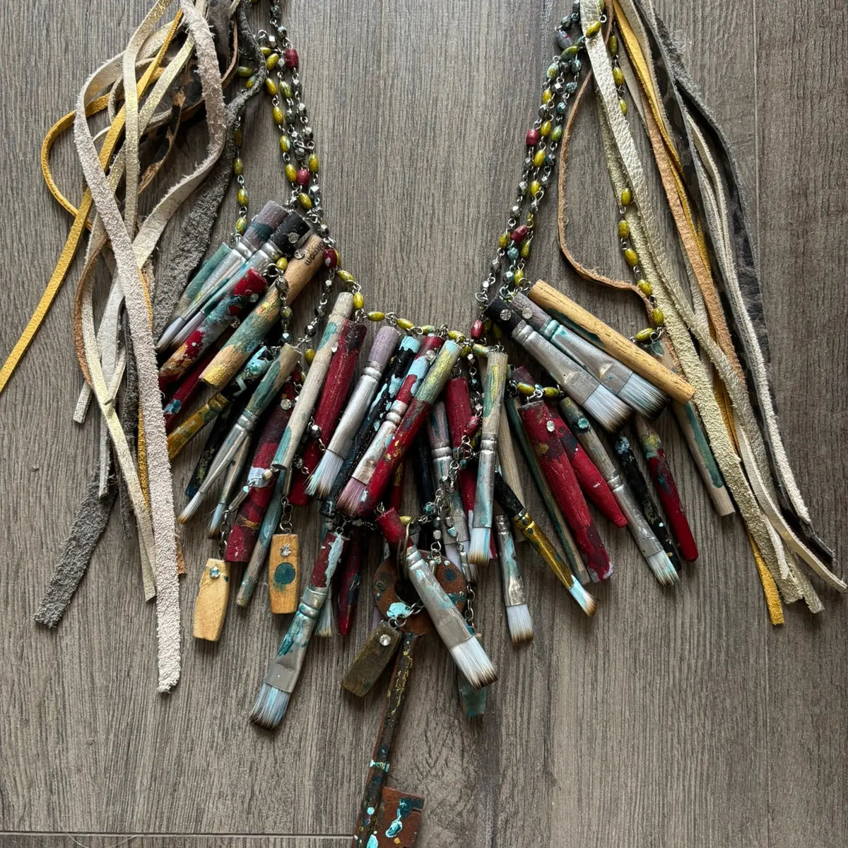 A Rare Bird Painter's Palette Layered Necklace, a wide bib-style statement necklace with layered paint-marked brushes, colorful beads, chains, dangling artist-inspired objects, and long mixed leather fringe ties.
