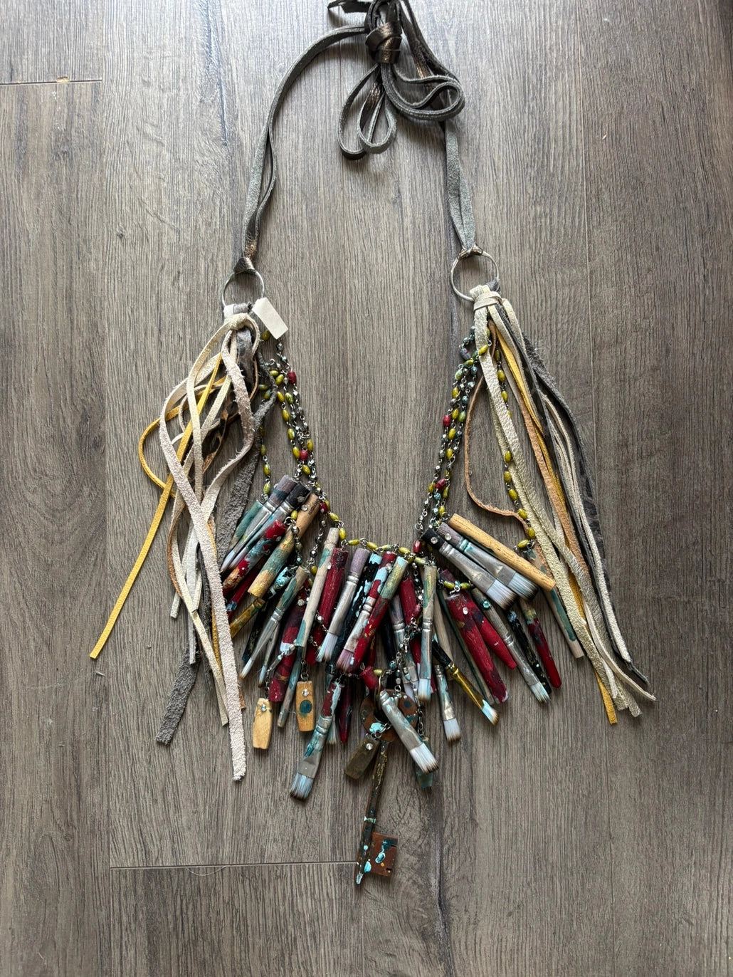 A Rare Bird Painter's Palette Layered Necklace, a wide bib-style statement necklace with layered paint-marked brushes, colorful beads, chains, dangling artist-inspired objects, and long mixed leather fringe ties.